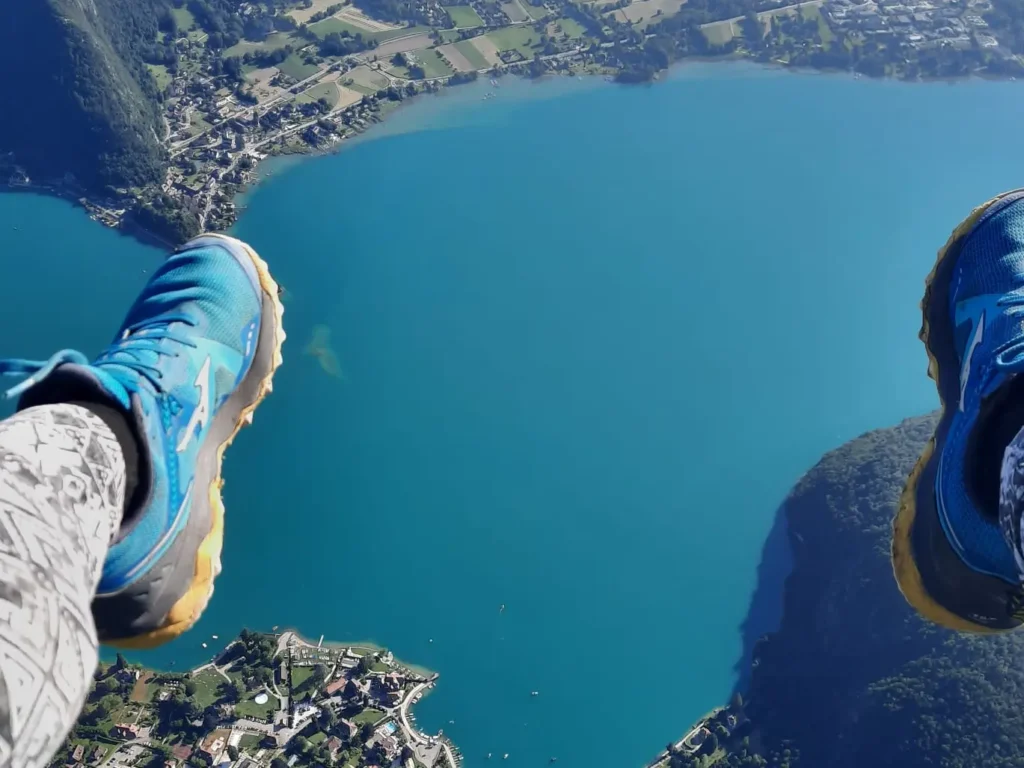 Les pieds d'un passagers en parapente sont au premier plan, en arrière plan, le lac Annecy tout petit et sa côte avec maisons et montagne