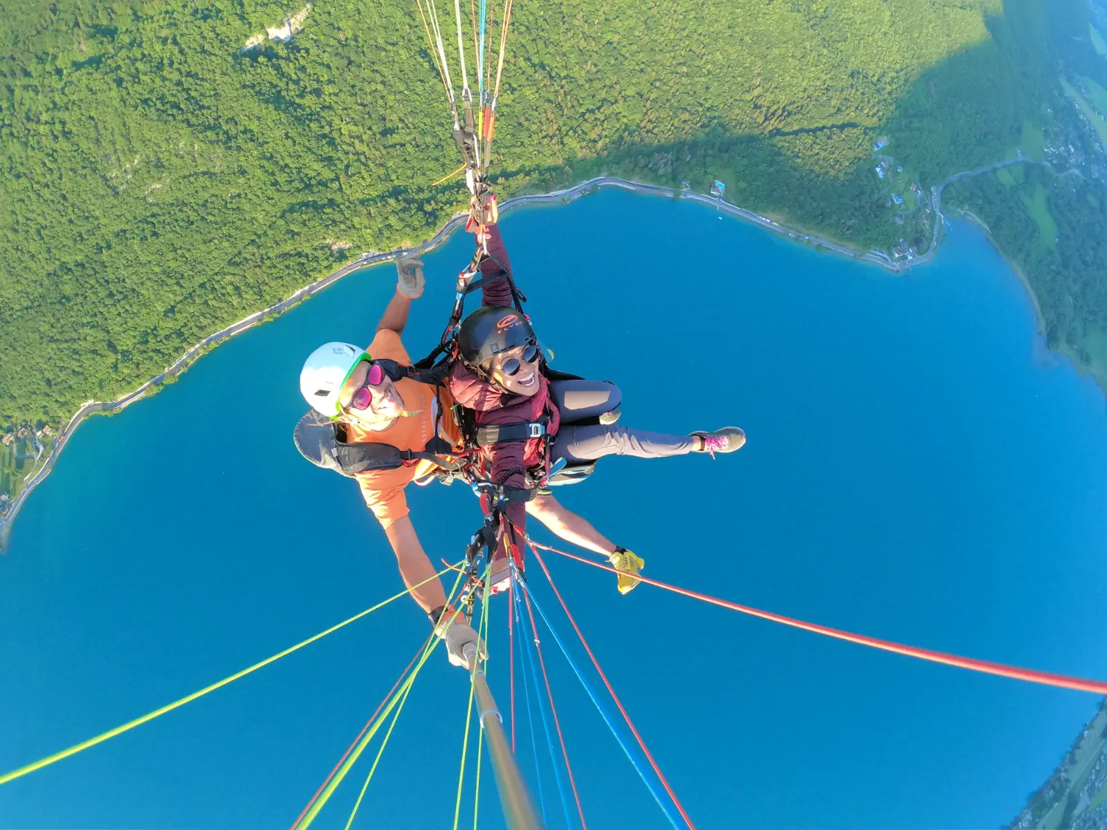 A paragliding pilot and his passenger are seen from above with the paraglider lines that attach them to the sail and Lake Annecy below