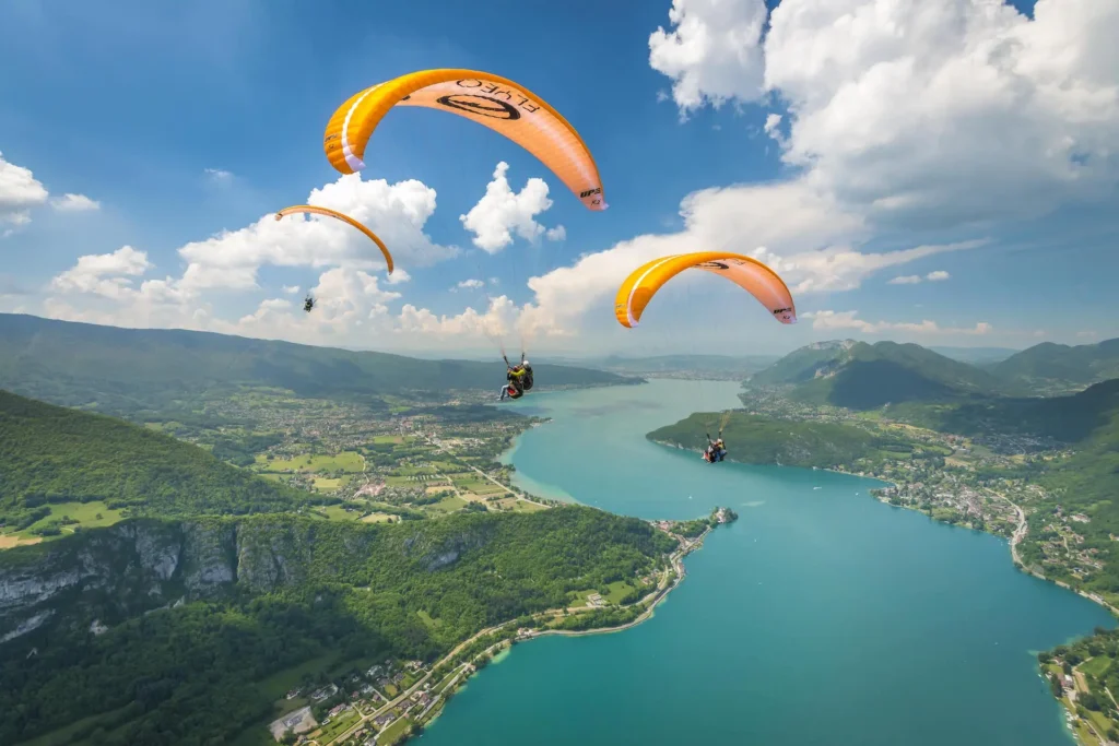 Two orange Flyeo paragliders fly over the blue waters of lake annecy
