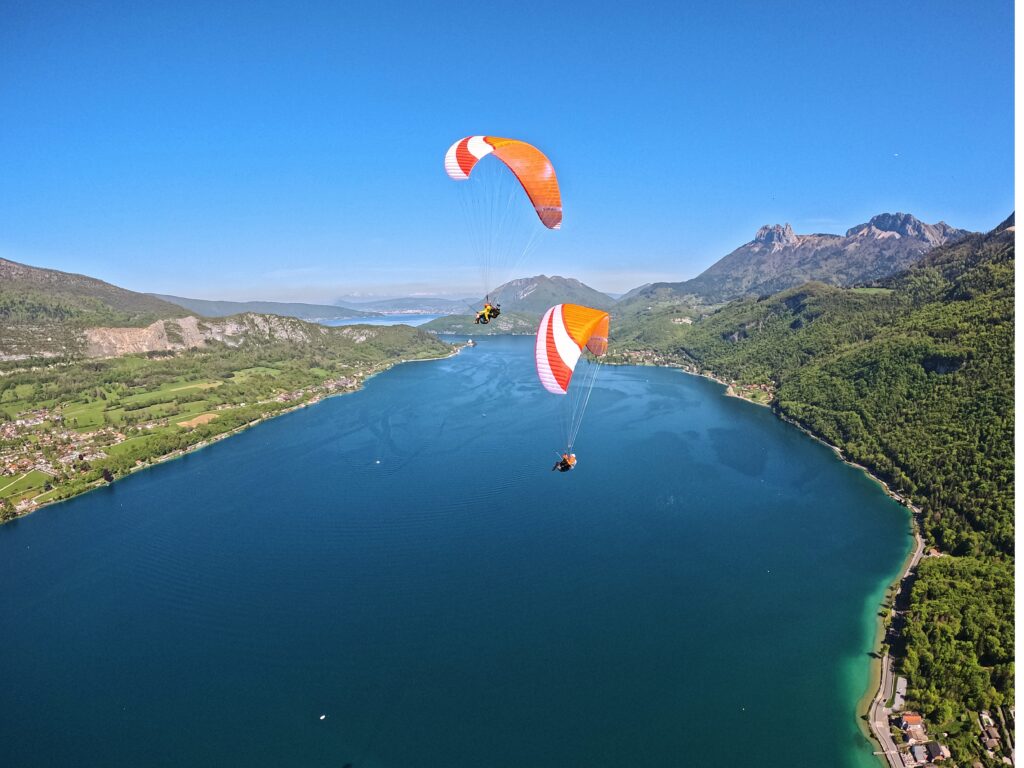 Deux parapentes biplaces orange Flyeo volent en formation au dessus du milieu du lac Annecy aux couleurs bleues complexes
