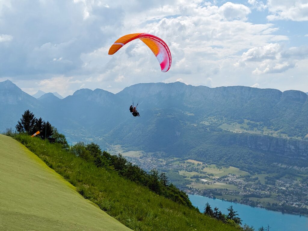 Un pilote et son passager sous leur parapente orange viennent juste de décoller. En fond, le lac d'Annecy et en premier plan, le tapis vert du décollage