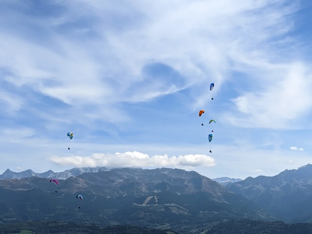 Cinq parapentes sont vus en vol avec en une chaine de montagnes et des nuages en fond